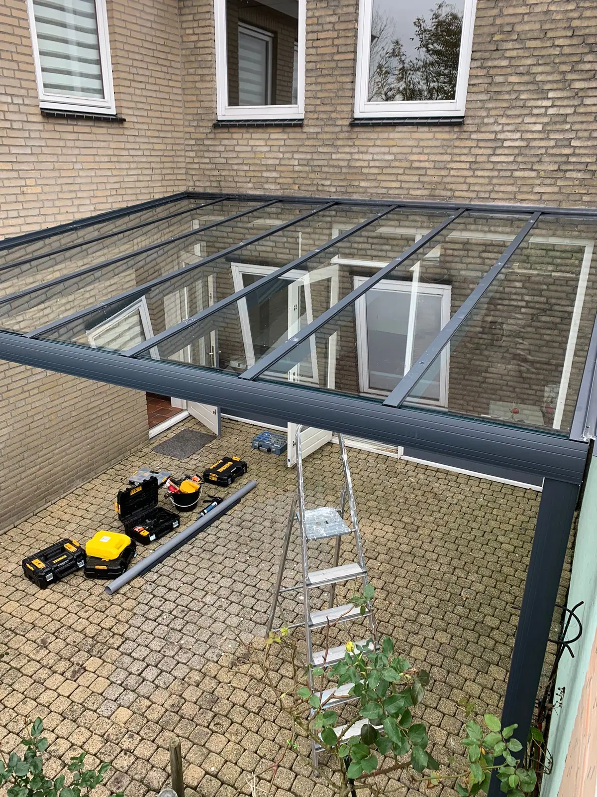 Een glazen dak van bovenaf gezien, met gereedschapskoffers en een ladder op de bestrating eronder